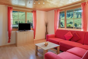 A room with a wooden ceiling, a television on a cabinet, a red sofa, and a view of a green landscape through large windows. - Bühlhof Todtnauberg Apartment 3 (Todtnau)