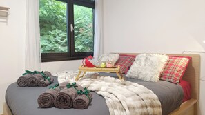 Ein Schlafzimmer mit Bett, einem Fenster mit Blick auf Grünflächen, einem Nachttisch mit rotem Teekessel und gerollten Handtüchern auf dem Bett.