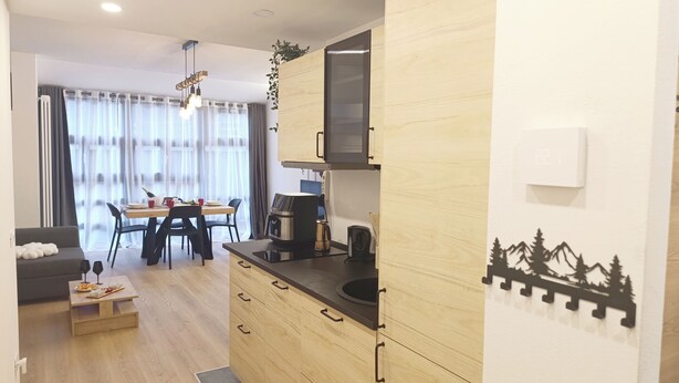 A modern kitchen with wooden cabinets, a dining area with a table and chairs, and a living area with a sofa and coffee table. - Apricola By ca-sa check (Aprica)