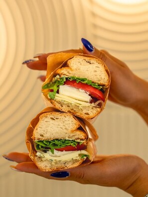 A sandwich cut in half, held in a paper wrapper, with visible ingredients like lettuce, tomato, and cheese. - Luz Suit (Kaş)