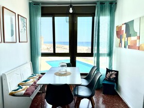 A dining area with a table and chairs, a view of the beach, and a window with curtains. - Sea View Apartment with Pool in Almería for up to 4 Guests (Retamar)