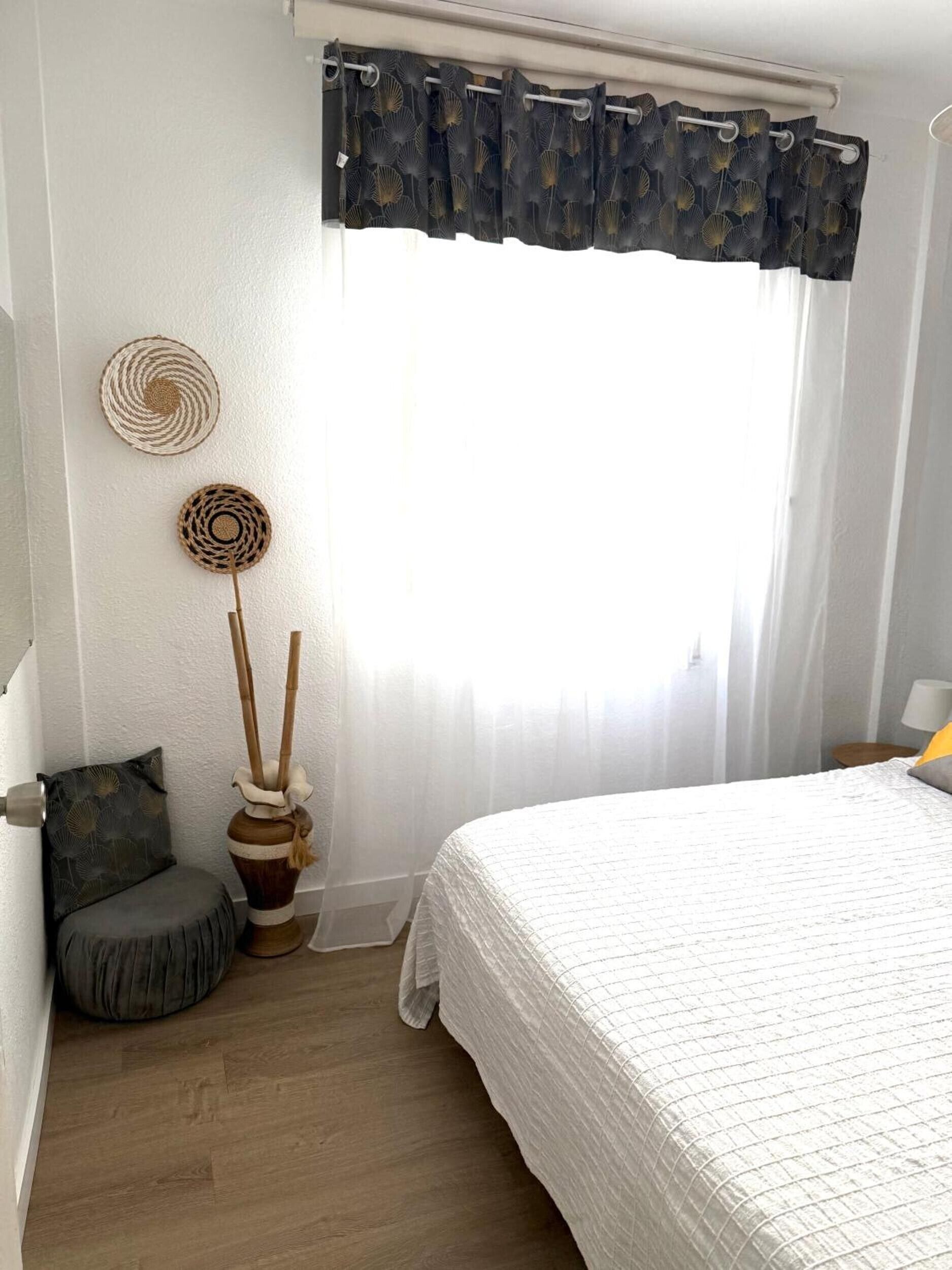 Un dormitorio con cama, una ventana con cortinas translúcidas, una mesita de noche con lámpara y un adorno en la pared.