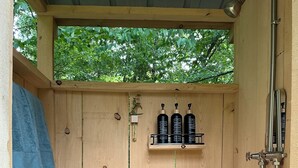 A wooden shower enclosure with a shelf holding shampoo, conditioner, and body wash bottles, a showerhead, and a towel hanging on the left. - The Woodland Retreat - Private Glamping Under Dark Skies (Long Eddy)