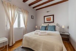 Ein Schlafzimmer mit einem Bett, Nachttischen, einem Fenster mit Vorhängen, einem gerahmten Bild an der Wand und Holzbalken an der Decke.
