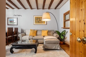Ein Wohnzimmer mit einer Couch, Hocker, Couchtisch, Holzstühlen, einer Lampe, einem gerahmten Bild und einer Tür.
