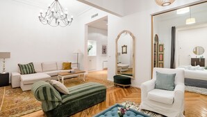 A well-lit living room with a chandelier, a sofa, a chair, a mirror, and a wooden floor. - Waou Gran Via IX - Short-Term Rental (Madrid)