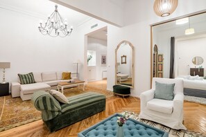 Ein helles Wohnzimmer mit einer Chandelüster, einem Sofa, einem Sessel, einem Spiegel und einem Holzboden.