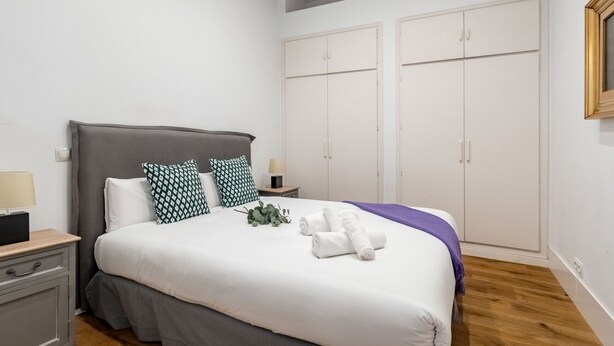 A neatly made bed with white linens, a grey headboard, and two nightstands with lamps. - Waou Gran Via IX - Short-Term Rental (Madrid)