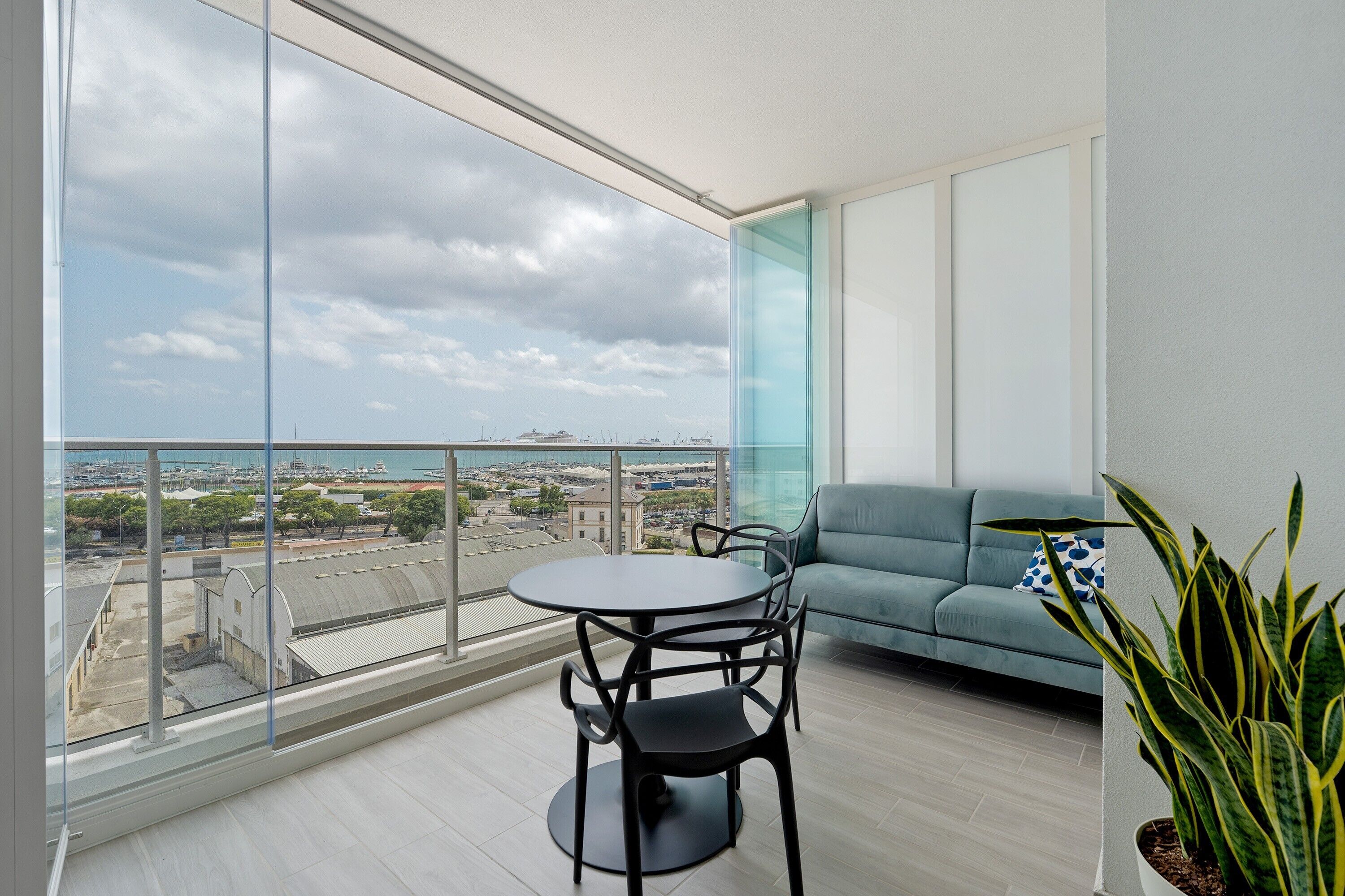 Un balcone con vista sul porto turistico e sulla città, un tavolo rotondo con sedie e un divano azzurro chiaro.