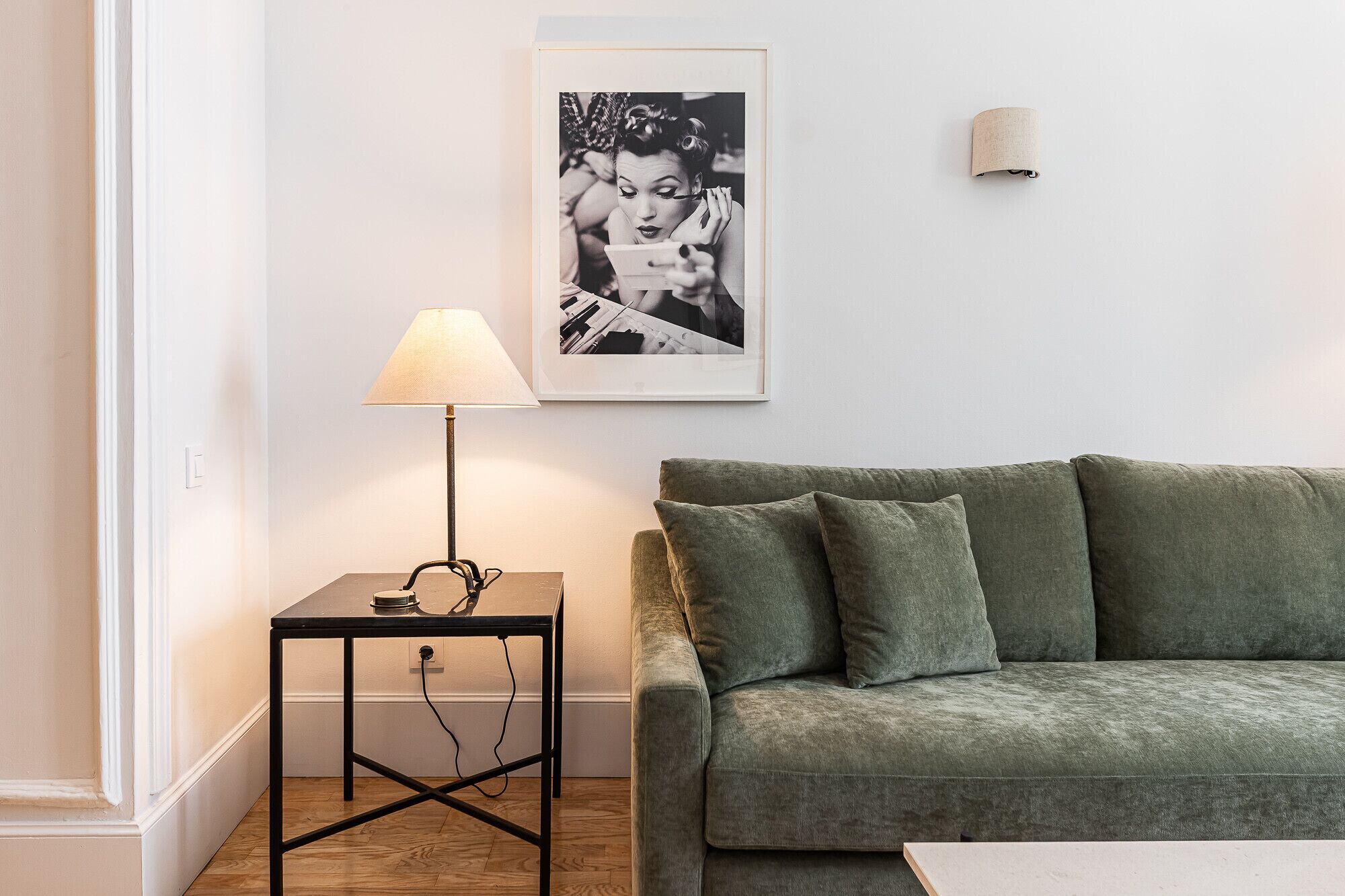 Ett vardagsrum med en grön soffa, ett litet bord med en lampa och en inramad tavla på väggen.