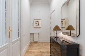 A hallway with a white door, a framed artwork on the wall, a radiator, and a mirror with two lamps. - Waou Sol XI - Short-Term Rental (Madrid)