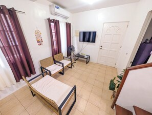 Una habitación con cama, dos sillas, televisor, aire acondicionado, ventilador y una puerta.