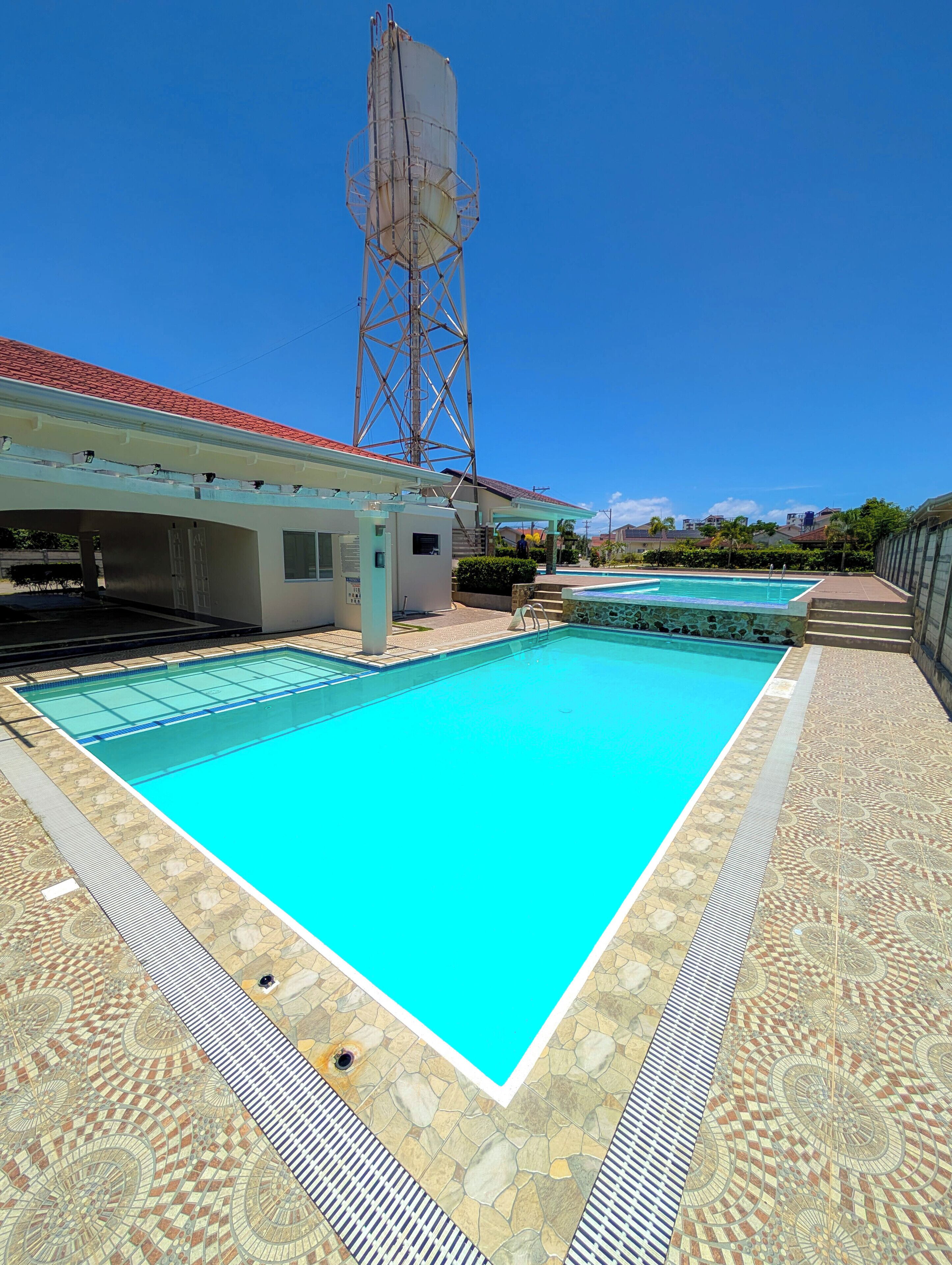 A swimming pool with a tiled deck, a covered area with a red roof, and a tall structure in the background.