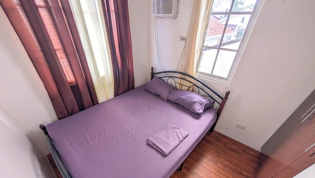 Un lit simple avec un cadre en métal, une fenêtre avec des rideaux et un plancher en bois.