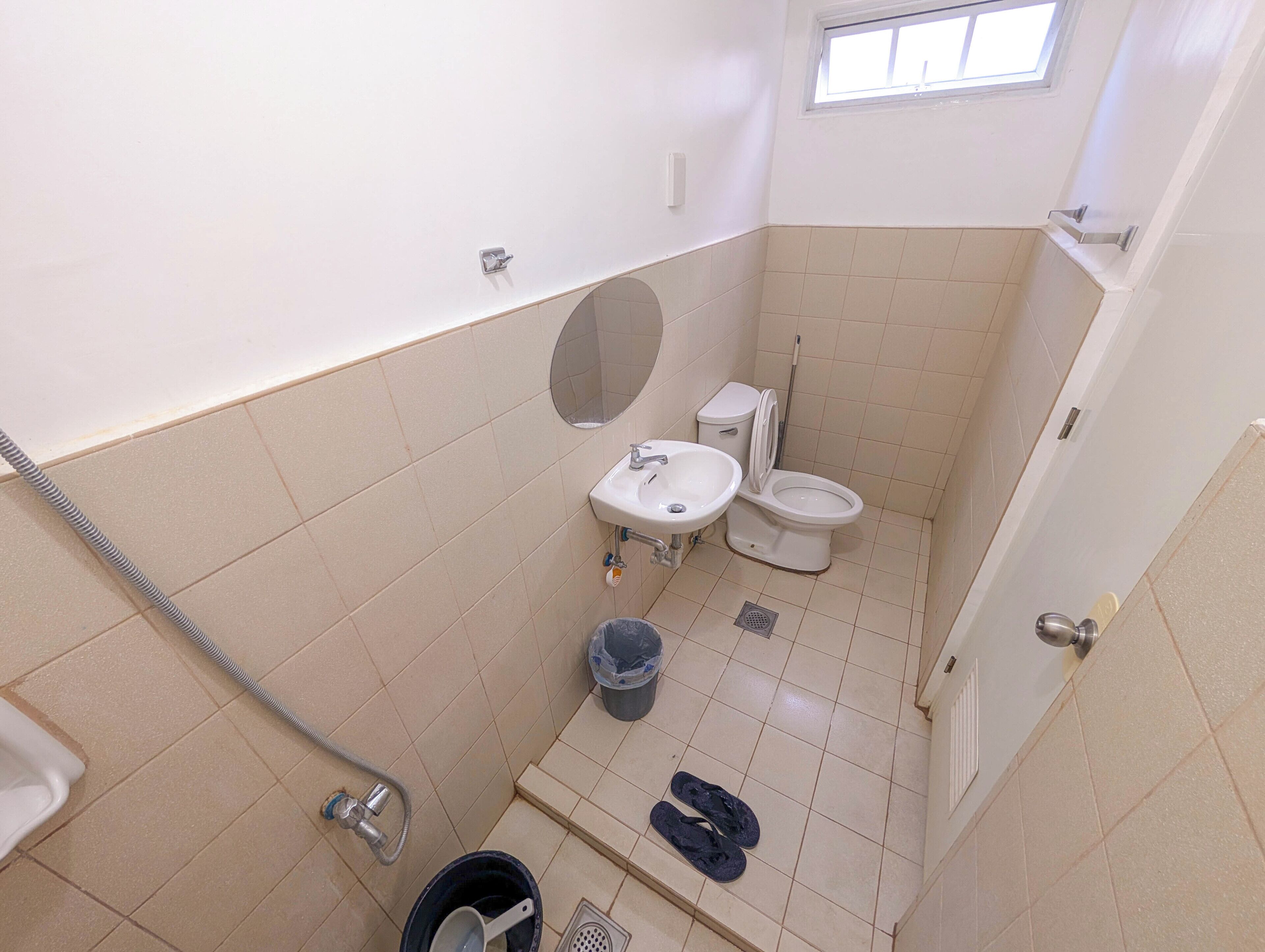 Ein kleines Badezimmer mit Toilette, Waschbecken und Dusche. Über der Toilette befindet sich ein Fenster und rechts eine Tür.