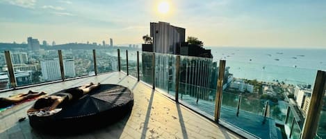A balcony with a view of the city and the sea, a black lounge chair, and a glass railing.