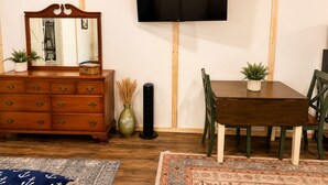 Um quarto com cômoda de madeira, espelho, televisão de tela plana na parede, mesa de jantar com cadeiras e um tapete estampado no chão.