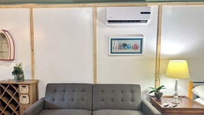Um sofá cinzento, uma mesinha com abajur, um ar condicionado fixado na parede, um quadro emoldurado e um armário de madeira com cestos.