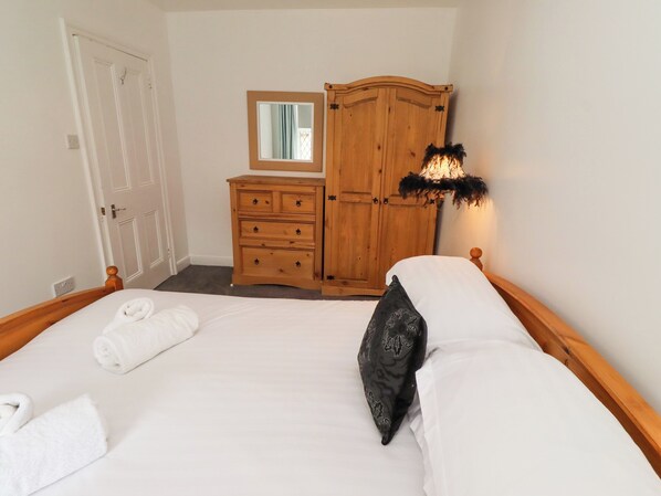 A bedroom with a bed, a wooden dresser, a mirror, and a wall-mounted lamp. - Beacon Cottage (Seahouses)