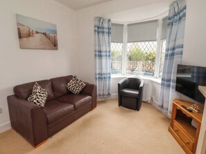 A living room with a brown leather sofa, a black armchair, a television on a wooden stand, and a window with blue curtains. - Beacon Cottage (Seahouses)