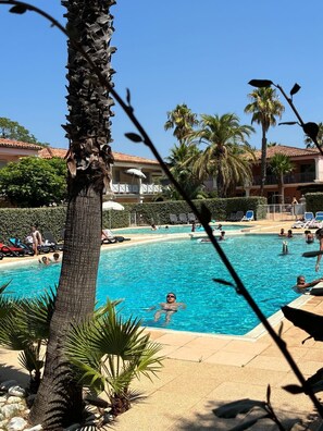 Ein Swimmingpool mit darin badenden Personen, umgeben von Palmen und einem Gebäude mit einem Ziegeldach.