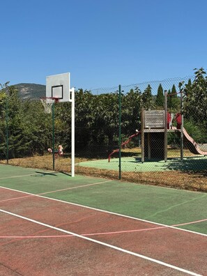 Ein Basketballfeld mit Korb, eine Rutsche und ein eingezäunter Bereich mit Bäumen und einer hügeligen Landschaft im Hintergrund.