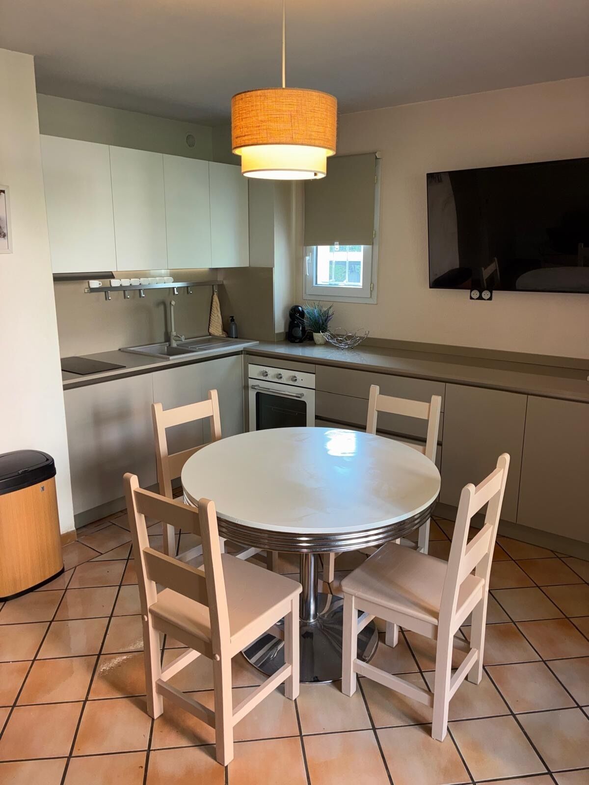 A modern kitchen with a dining table and chairs, a hanging light fixture, and a flat-screen TV mounted on the wall.