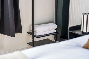 A hotel room with a bed, a shelf with folded towels, a black coat hanging, and a black trash bin. - Comfy Studio for Solo Travellers| Free Parking|CV2 (Coventry)