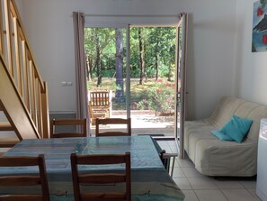 Une pièce comprenant une table à manger, des chaises, un canapé et une porte-fenêtre coulissante donnant sur un espace extérieur agrémenté de verdure et délimité par une clôture.