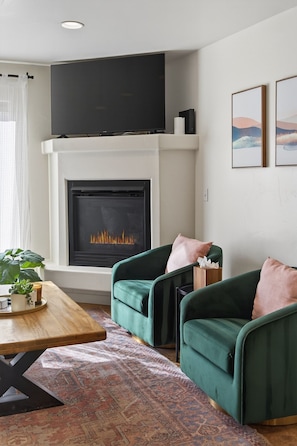 A living room with a fireplace, two green armchairs, a wooden coffee table, and a patterned rug. - Modern Rim Village Retreat- Pool, Hot Tub, Red Rock, and Mountain Views. (Moab)