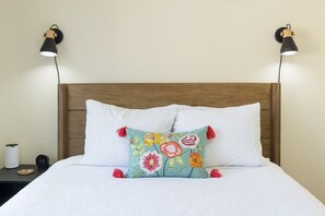 Ein Bett mit weißer Bettwäsche, einem farbenfrohen Blumenkissen und zwei Wandlampen.