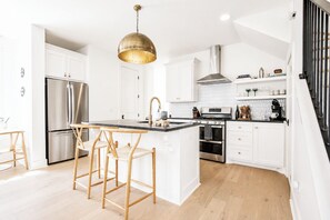 Eine moderne Küche mit weißen Schränken, Edelstahlgeräten, einer zentralen Kochinsel mit Hockern und einer hängenden Pendelleuchte.
