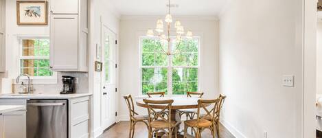 A kitchen with white cabinetry, a stainless steel dishwasher, and a dining area with a wooden table and chairs.