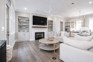 A modern living room with a fireplace, a flat-screen TV, a ceiling fan, and a kitchen area with a stainless steel range hood. - The Social, 3mi to SQ + Pool by VelvetDitchVillas (Oxford)