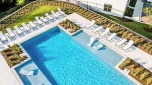 En rektangulär swimmingpool med ett klart, blått vatten, omgiven av en stenlagd terrass med vita solstolar och små, välskötta trädgårdsområden.