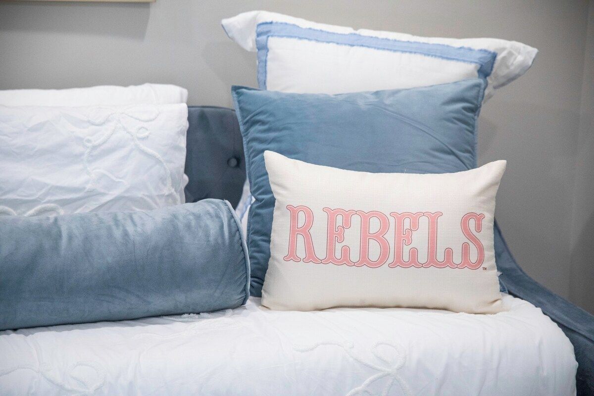 A bed with multiple pillows in shades of blue and white, one pillow with the word 'REBELS' embroidered on it.