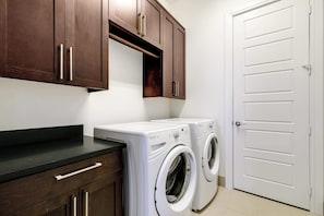 A laundry room with two white front-loading washing machines, wooden cabinets, and a white door. - Luxury 3BR Penthouse |Skyline Views |Uptown Dallas (Dallas)