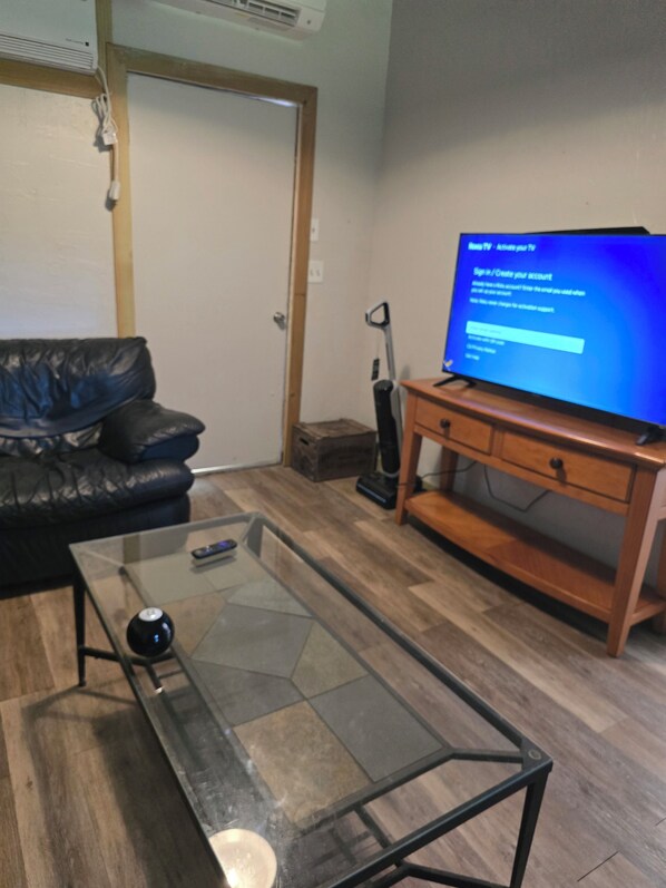 A living room with a glass-top coffee table, a black leather sofa, a wooden TV stand, and a vacuum cleaner.