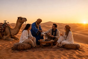 Un grupo de personas sentadas sobre una alfombra en un paisaje desértico, con camellos y una mesa con bebidas.