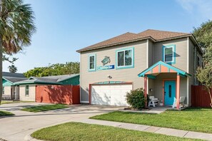 A two-story house with a garage, a covered porch with chairs, and a small garden area. - Heated Pool-Fun Game Room -Close to Beach-Pet Friendly (Galveston)