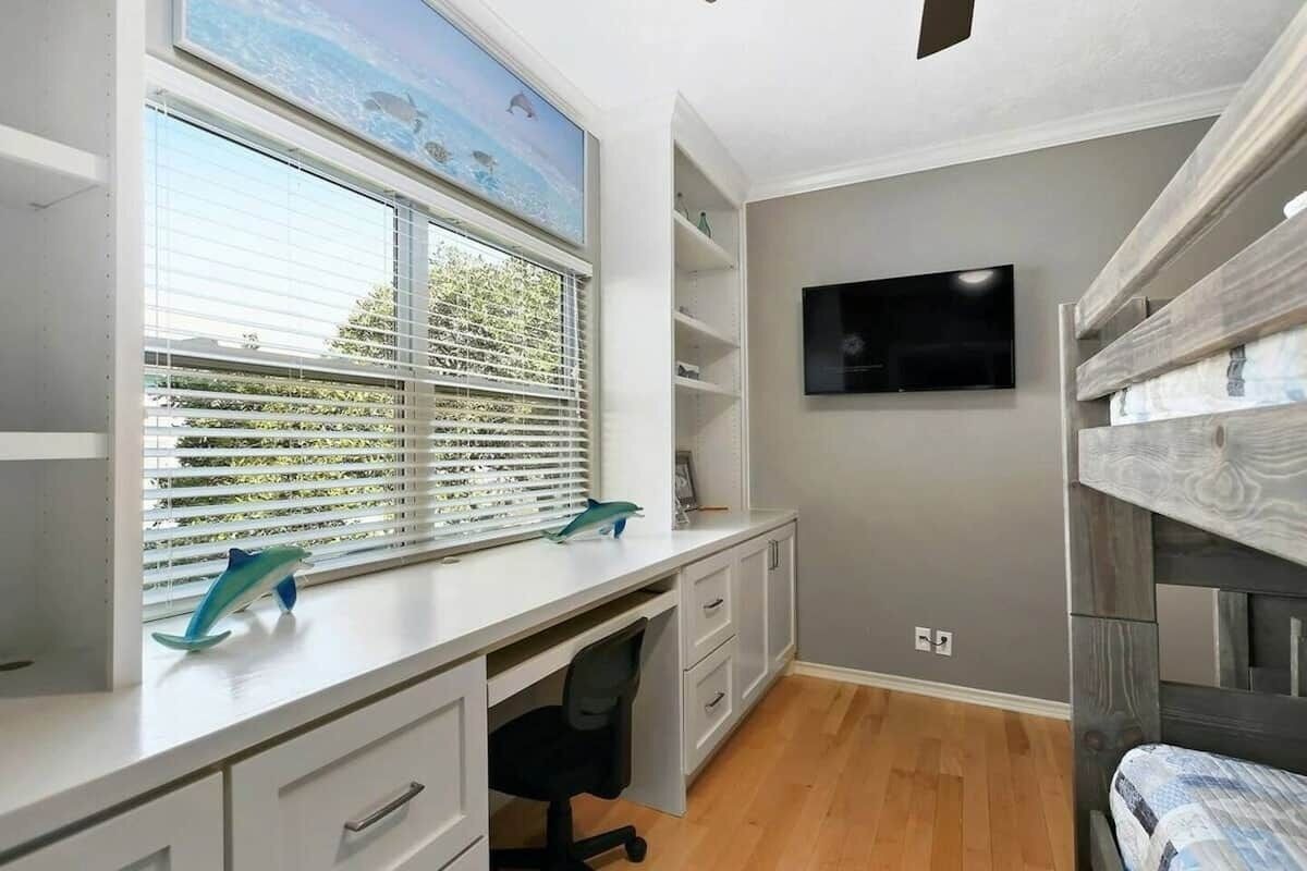 Un dormitorio con una cama litera, un escritorio con una silla, un televisor en la pared y una ventana con persianas.