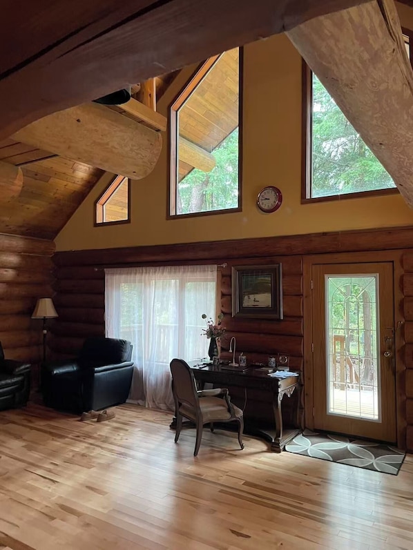 A log cabin interior with a wooden floor, a sofa, a desk, and a large window with a view of trees.