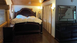 A bedroom with a wooden ceiling, a bed with white bedding, a nightstand, a lamp, a wardrobe, and a mirror.