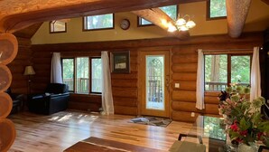 A log cabin interior with a wooden floor, a sofa, a dining table with chairs, and a large window with curtains.
