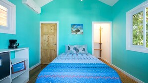 A room with teal walls, a wooden door, a bed with patterned bedding, a shelf with a coffee maker, and a framed picture on the wall.