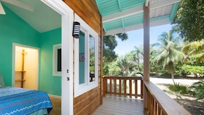 A room with a bed, a door, and a wooden balcony with a view of palm trees.