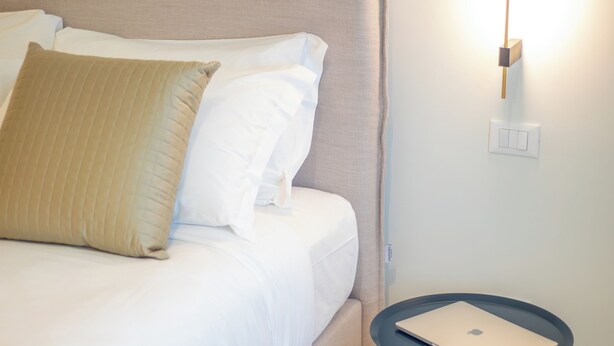 Una cama bien hecha con sábanas blancas y un cabecero beige, una mesita de noche con un ordenador portátil, y una lámpara de pared.