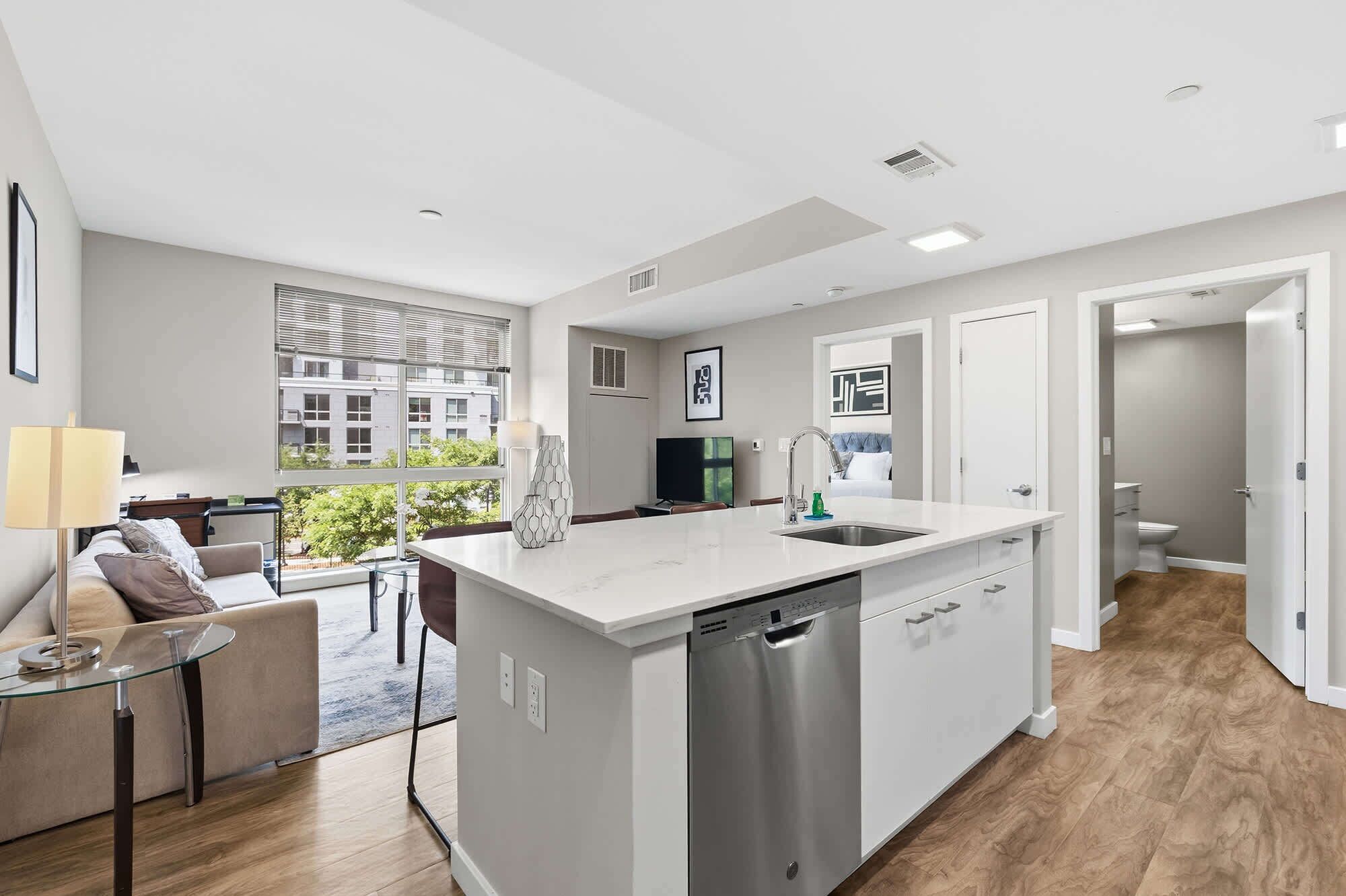 A modern kitchen with a dishwasher, a dining area with a glass table, and a view of the cityscape.