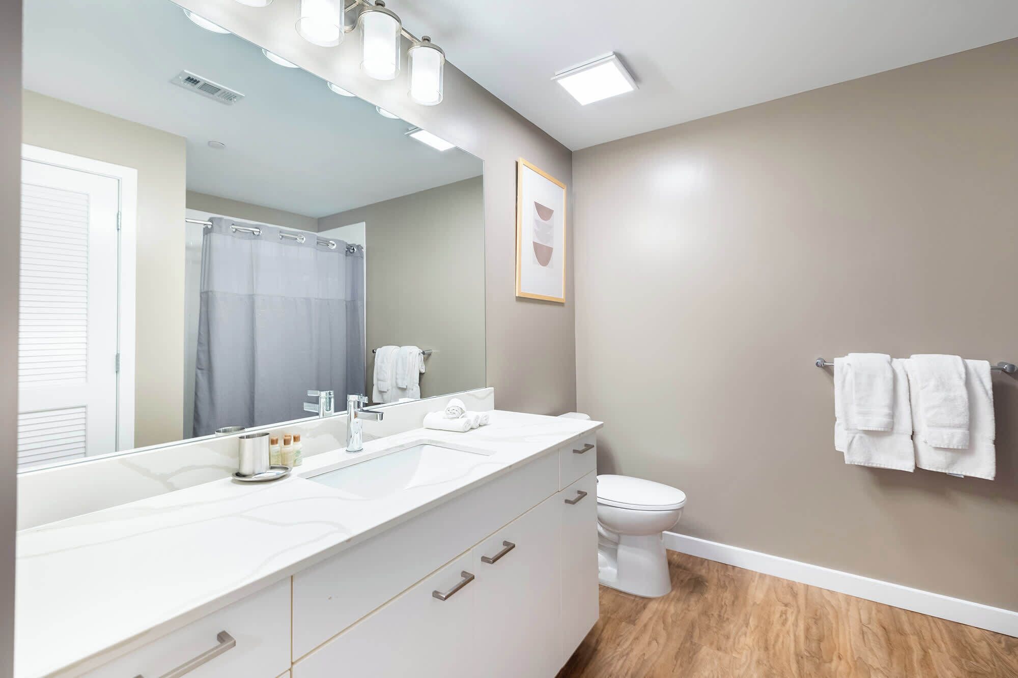 A modern bathroom with a large mirror, a shower, a toilet, and a vanity with drawers.