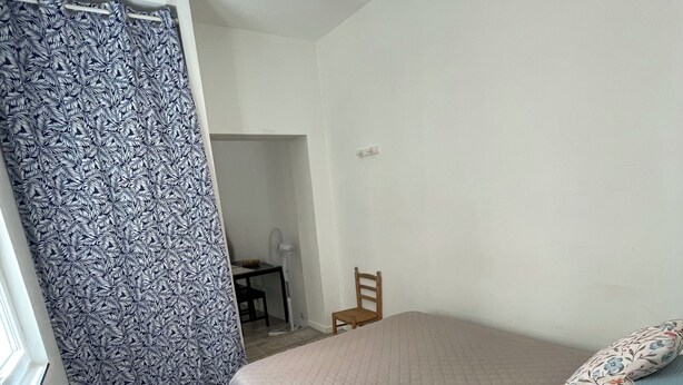 Ein Schlafzimmer mit einem Bett, einem Sessel, einem Schreibtisch und einem gemusterten Vorhang.